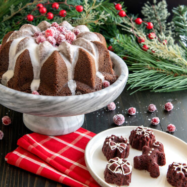 Chocolate bundt cake with white glaze on cake stand, baked in Nordic Ware Vintage Star pan, topped with cranberries