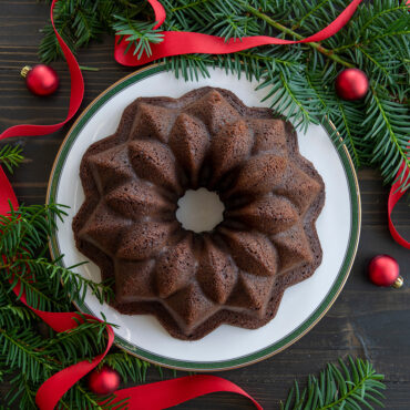 Chocolate bundt cake with star-shaped ridged design on white plate, surrounded by evergreen sprigs and red ribbon
