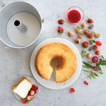 Plain white angel food cake on plate with fresh strawberries, Nordic Ware aluminum angel food pan with removable cone, and slice on small plate