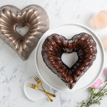 Elegant Heart Bundt cake with chocolate frosting on white cake stand, surrounded by pink roses and pan, overhead