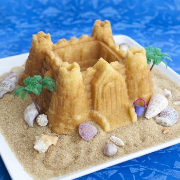 Golden castle-shaped bundt cake on beach-themed display with brown sugar sand, seashells and palm trees