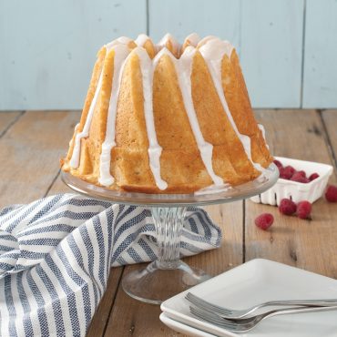 Golden kugelhopf bundt cake with white glaze drizzle on glass cake stand, fresh raspberries alongside