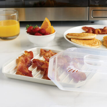 Nordic Ware white microwave bacon tray with clear lid, containing crispy cooked bacon strips on ridged surface