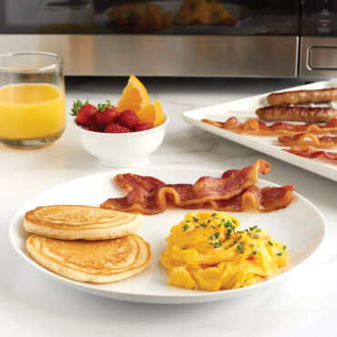 Nordic Ware large slanted aluminum bacon tray with cooked bacon strips, displayed with breakfast spread