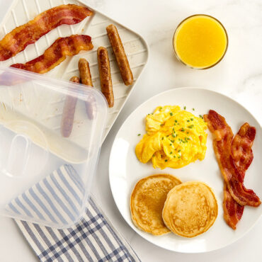 Nordic Ware gray aluminum slanted bacon tray with lid, containing cooked bacon strips and breakfast sausages