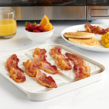 Nordic Ware medium slanted bacon tray with clear lid, containing cooked bacon strips on kitchen counter with breakfast spread