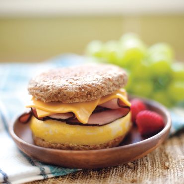 Breakfast sandwich with melted cheese, Canadian bacon, and cooked egg on whole grain English muffin served on brown plate