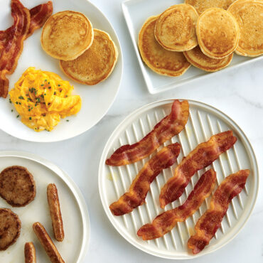 Two-sided aluminum meat grill with ribbed and flat surfaces, cooked bacon and sausages, served with plated breakfast items