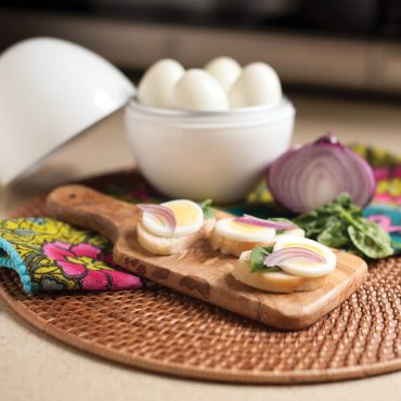 Nordic Ware egg boiler with perfectly sliced hard-boiled eggs arranged on wooden cutting board, garnished with red onion and fresh basil