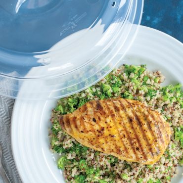 Nordic Ware clear microwave spatter cover beside white plate with grilled chicken breast and quinoa salad