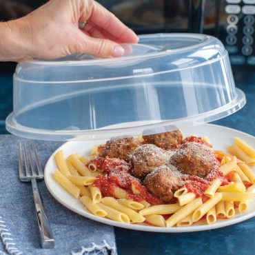 Clear plastic microwave spatter cover being lifted from white plate of penne pasta with meatballs and tomato sauce