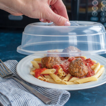 Hand holding clear plastic microwave spatter cover over plate of penne pasta with meatballs in tomato sauce