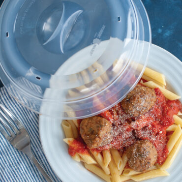 Clear spatter cover partially covering white plate of penne pasta with meatballs and red sauce