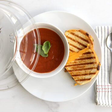 Nordic Ware clear glass deluxe plate cover with white plate, tomato soup bowl, grilled cheese sandwich