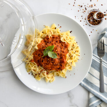 Clear Nordic Ware deluxe plate cover over white plate with spaghetti and red sauce, garnished with basil