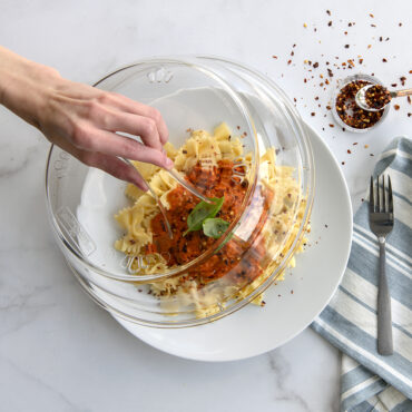 Nordic Ware clear microwave plate cover being lifted by hand, revealing pasta with marinara sauce and basil
