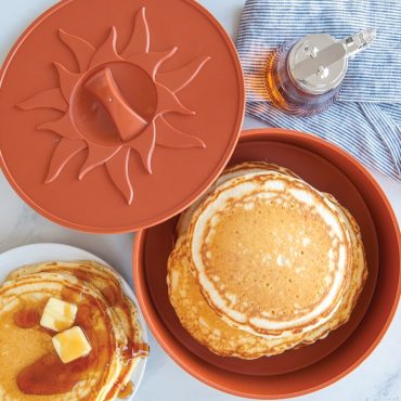 Nordic Ware orange tortilla warmer with sun design lid, stack of golden pancakes inside, syrup and butter nearby