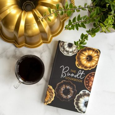 Nordic Ware Anniversary Bundt pan with The Bundt Cookbook, coffee cup and plant on white marble surface