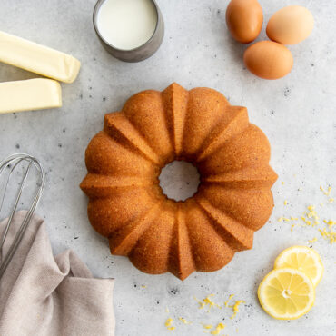 Golden lemon bundt cake in Nordic Ware fluted pan surrounded by fresh eggs, butter, milk and lemon