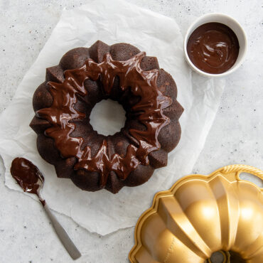Dark chocolate bundt cake with glossy chocolate glaze, golden Nordic Ware bundt pan beside it