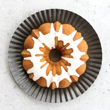 Glazed vanilla bundt cake on white plate, baked in Nordic Ware Original aluminum bundt pan with fluted design