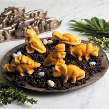 dinosaur cakelets on wooden plate with crushed cookies and candy dinosaur eggs with pan and green leaves