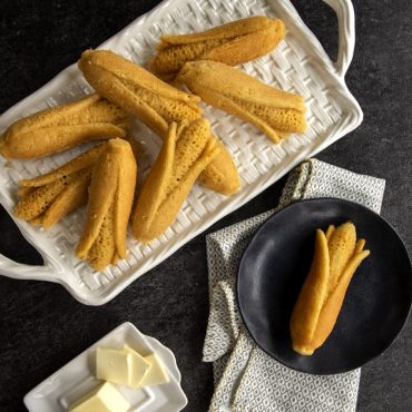 Golden cornbread cakes baked in Nordic Ware corn-shaped pan, displayed on slate platter with butter pat