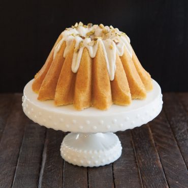 Golden bundt cake baked in Nordic Ware Lotus bundt pan with white glaze dripping down fluted sides, topped with chopped nuts on white cake stand