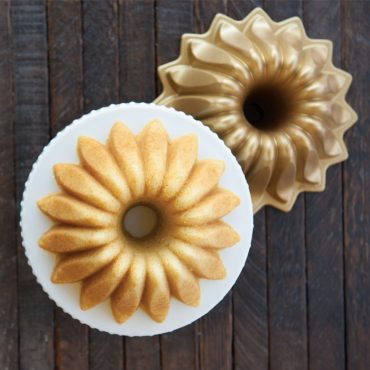 Golden vanilla bundt cake with lotus petal design on white plate beside Nordic Ware aluminum Lotus bundt pan