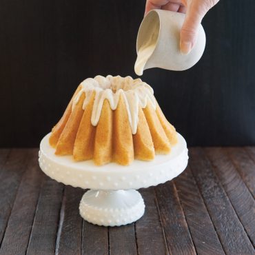 Golden bundt cake with lotus petal design on white cake stand, white glaze being poured from small pitcher