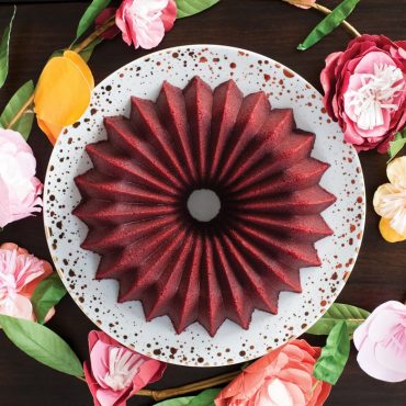 Red velvet bundt cake with smooth glaze on white plate, surrounded by pink and white flowers