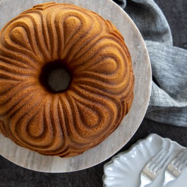 Golden brown chiffon bundt cake with decorative swirled ridges on white cake stand, baked in Nordic Ware pan