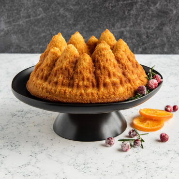 Golden bundt cake with detailed pine tree design on white stand, garnished with orange slice and berries