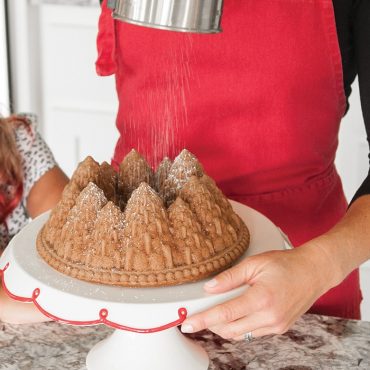Woman dusting powdered sugar on golden bundt cake with detailed pine tree design from Nordic Ware Pine Forest pan