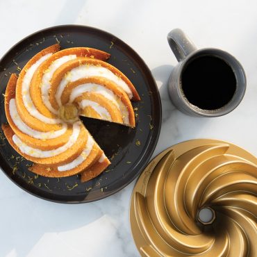Golden bundt cake with white glaze on dark plate, Nordic Ware Heritage aluminum bundt pan and coffee cup nearby