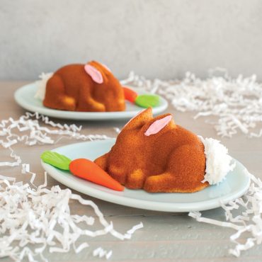 Two golden vanilla bunny cakes from Nordic Ware pan with pink frosting ears, coconut tails, candy carrots