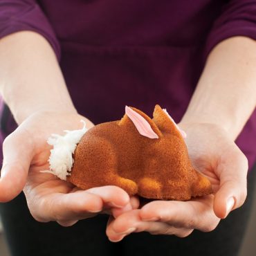 Hands holding golden-brown bunny-shaped cakelet with white frosting details, baked in Nordic Ware aluminum mold