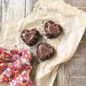 Three chocolate heart cakelets with glossy glaze and sprinkles on parchment paper, baked in tiered design