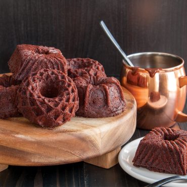 Six chocolate bundt cakes with geometric ridged patterns on wooden serving board, copper pot with spoon nearby