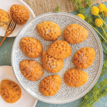 Six golden egg-shaped cakelets with ornate floral designs on white plates, blue background with fresh flowers