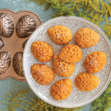 Six golden ornamental egg-shaped cakes on white plate, Nordic Ware aluminum cakelet pan visible in background