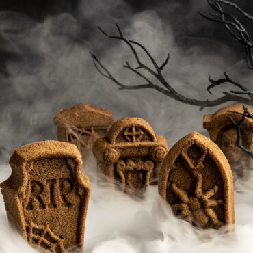 Golden baked tombstone-shaped cakes with detailed engravings, surrounded by spooky smoke and bare branches