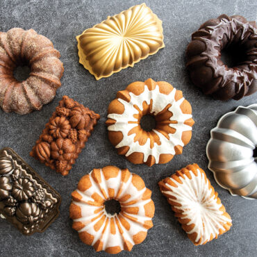 Multiple glazed bundt cakes and loaf cakes displayed with matching Nordic Ware aluminum baking pans on gray surface
