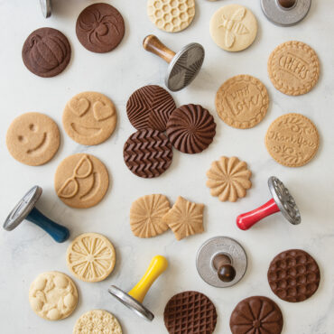 Six round baked cookies with geometric patterns next to Nordic Ware aluminum cookie stamps showing star, flower, and diamond designs