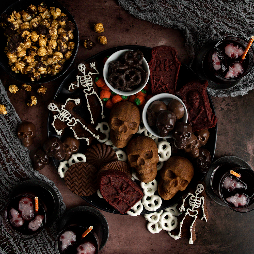 Halloween dessert board with skeleton-shaped cakes, orange treats, and spooky snacks arranged on dark surface