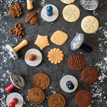 Holiday cookie stamps with baked cookies showing embossed winter designs, arranged on floured dark surface