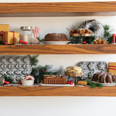 Nordic Ware bundt cakes and baking pans displayed on wooden shelves with holiday decorations and greenery