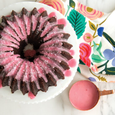 Chocolate bundt cake with pink glaze made in Nordic Ware 5-cup Brilliance pan, bowl of glaze beside floral towel