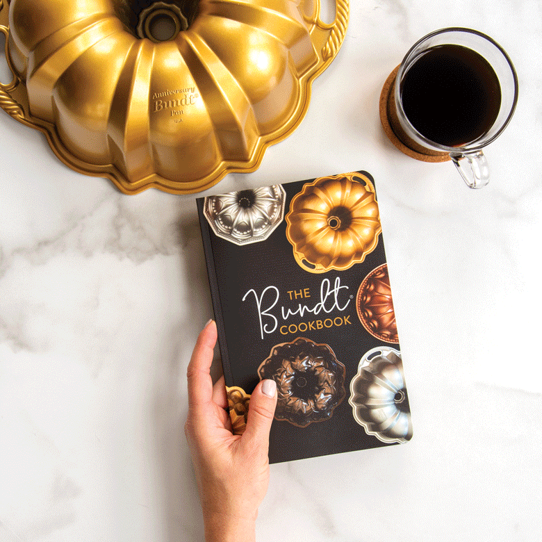 Nordic Ware Bundt Cookbook displayed on marble surface, hands flipping through pages showing recipes and bundt cake photos