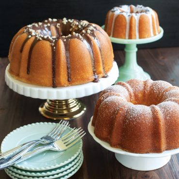 Three golden bundt cakes in graduated sizes on white cake stands, featuring chocolate drizzle, white glaze, and powdered sugar